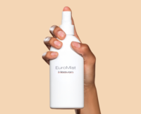 A hand holding a white spray bottle labeled EuroMist Heaven against a plain beige background.