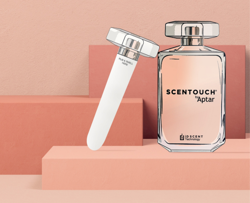 A clear perfume bottle labeled SCENTOUCH by Aptar stands next to a white reusable paper fragrance sampling applicator on pink geometric blocks, set against a peach background.