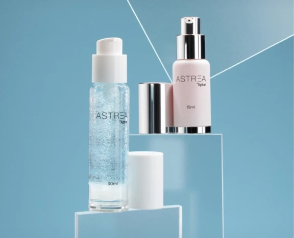 Two ASTREA airless packaging bottles —one clear with a pump (30ml), one opaque pink with a silver dispenser (15ml)—are displayed on clear podiums against a blue background, both with their lids placed nearby.
