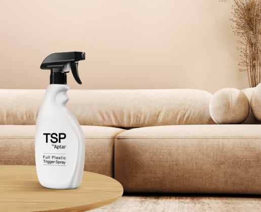 A white spray bottle labeled TSP Aptar Full-Plastic Trigger Spray Pump sits on a wooden table in front of a beige sofa with round cushions in a softly lit, modern living room.