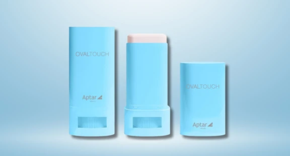 Two light blue Oval Touch on-the-go oval face stick containers labeled OVALTOUCH and Aptar beauty, standing upright against a soft blue gradient background. The first one is closed, middle container is open, revealing the white sick inside followed by the one cap.