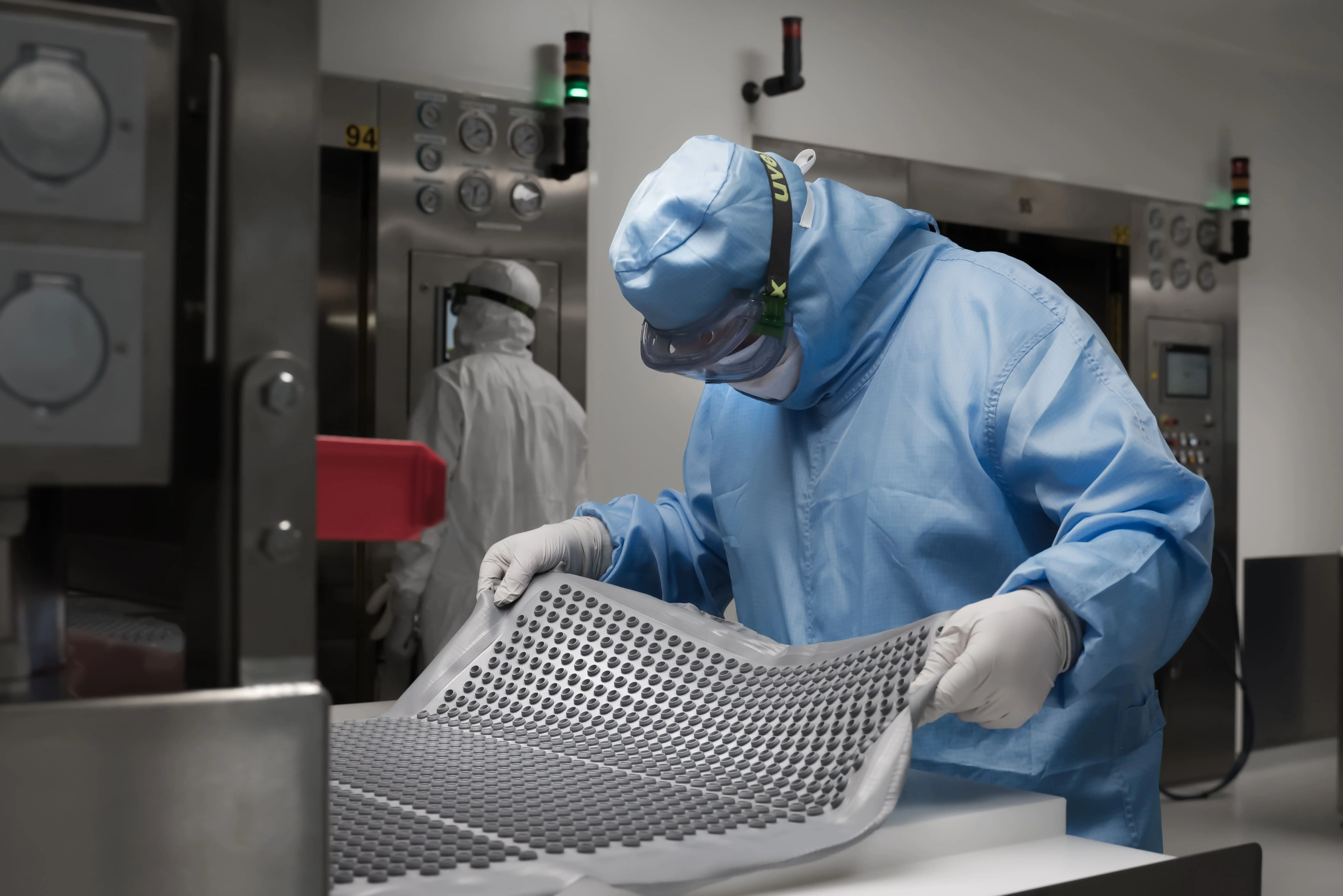 Cleanroom operator handling a tray of molded rubber stoppers in a controlled pharmaceutical manufacturing environment.