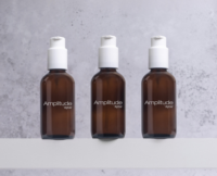 Three brown glass pump bottles labeled Amplitude Aptar, each featuring a high dose cosmetic pump, are placed side by side on a white surface against a light gray, textured background.