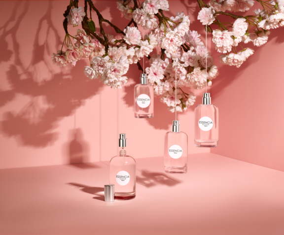 Four perfume bottles labeled Essencia are displayed in a pink setting, three gracefully hanging from cherry blossom branches and one upright with its elegant fragrance pump exposed. Pink blossoms fill the top of the image.