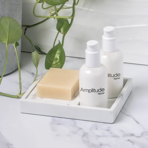 Two white bottles labeled Amplitude Aptar, a high dose cosmetic pump, and a beige bar of soap rest on a marble tray, with green leafy vines in the background atop a white, marble-like surface.