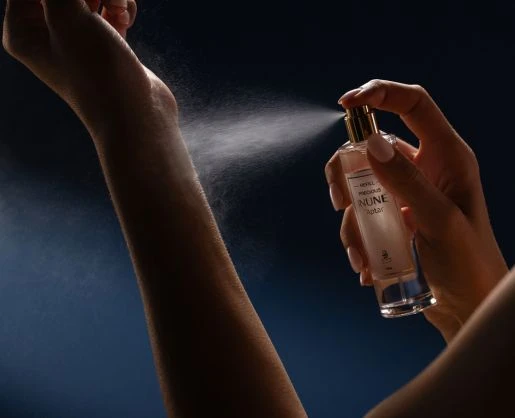 A hand holds an INUNE by Aptar spray bottle with a gold pump, releasing a fine mist toward an outstretched arm against a dark blue background.