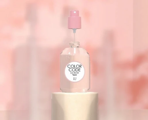 A clear glass bottle with a refillable plastic fragrance pump labeled Color Code Screw sits on a cylindrical pedestal against a soft pink, blurred background.