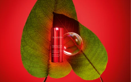 A red Clarins Total Eye Lift bottle, showcasing elegant beauty packaging, sits between two overlapping green leaves on a vibrant red background, accompanied by a clear spherical object.