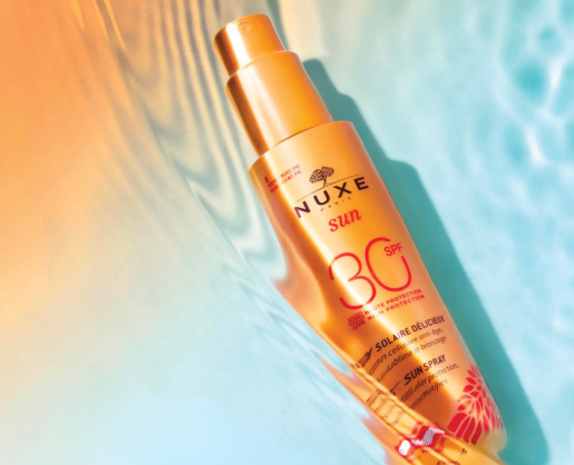 A gold bottle of Nuxe Sun SPF 30 sunscreen spray with featuring PZ Twist, Aptar Beauty's twist-to-lock-spray pump lies on a sunlit, rippled blue surface, creating a summery, vibrant atmosphere.