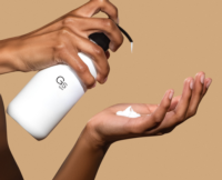 A person uses GS by Aptar ,an iconic high-performing dispensing pump to release lotion from a white bottle onto their palm, set against a beige background.