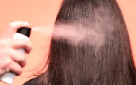 An image of a white aerosol spray can with a black actuator spraying dry shampoo and glitter on brown hair, depicting that a vertical slider aerosol valve is highly compatible with high powder formulations. The background is coral pink
