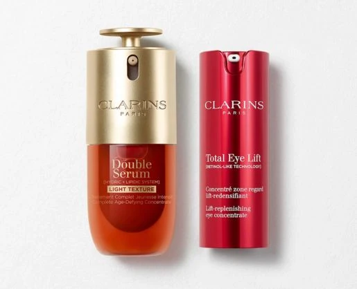 Two Clarins Paris skincare products, showcased in custom primary beauty packaging: Double Serum in a gold and orange bottle, and Total Eye Lift in a tall, red bottle-both laying flat on on a light textured surface.