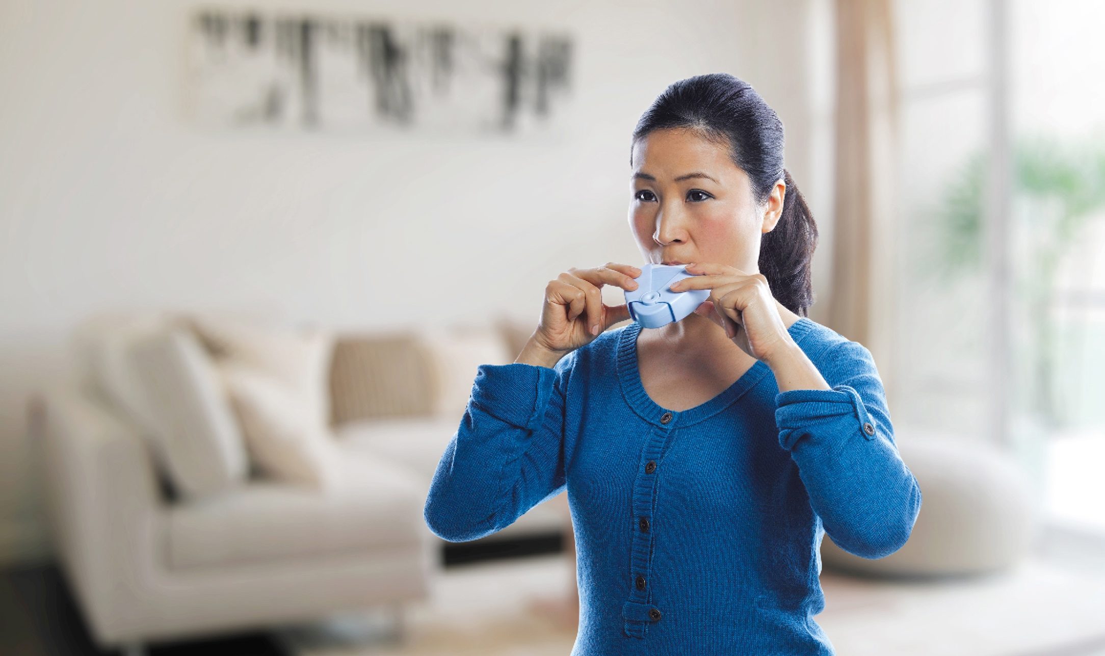 Woman with dark hair wearing blue sweater in her house is holding blue Aptar Pharma ProHaler® Inhaler solution to her mouth.
