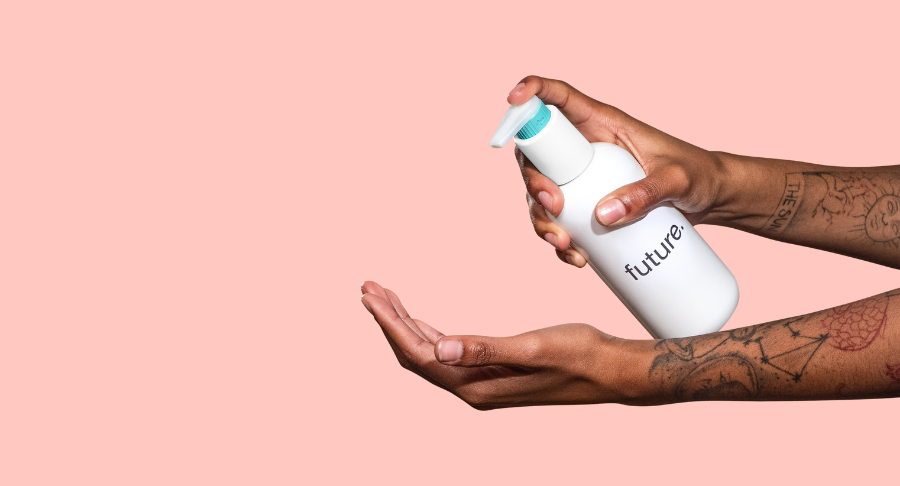 A person with tattooed arms dispenses lotion from a white pump bottle labeled future, a fully recyclable dispensing pump, onto their palm against a solid pink background.