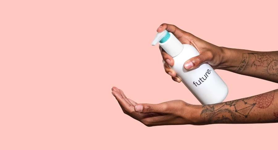 A person with tattooed arms dispenses lotion from a white pump bottle labeled future, a fully recyclable dispensing pump, onto their palm against a solid pink background.