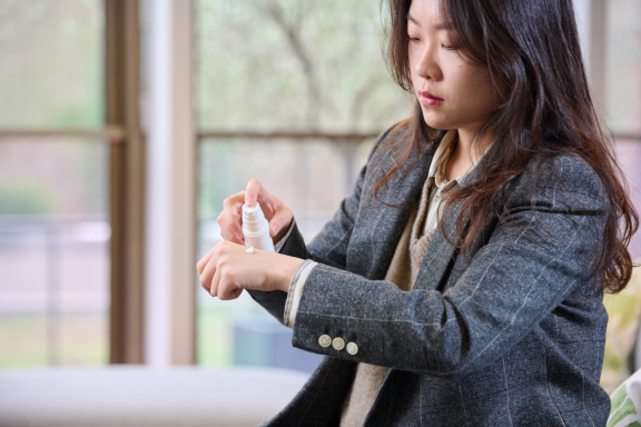 Woman with long dark hair wearing grey suit jacket dispenses dermal product on hand from white Aptar Airless+ delivery system