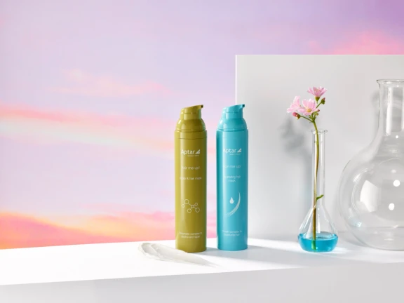 Two bottles of hair masks in airless packaging named Micro, one yellow-green and one blue, stand on a white surface with a glass vase in the shape of a beaker holding a pink flower and a round glass vase. The pastel pink and purple background resembles a soft sunset sky.