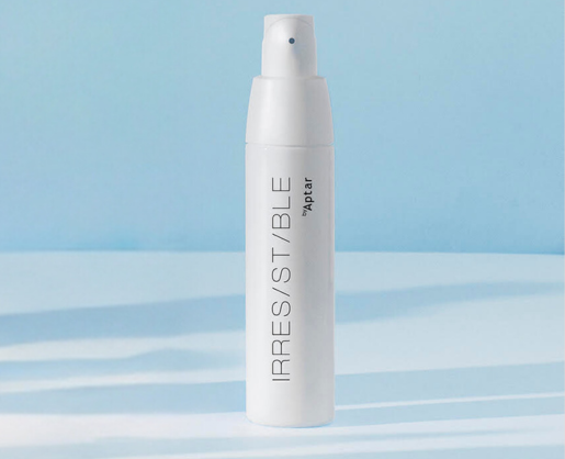 A white, bag-in-bottle airless pump bottle labeled IRRESISTIBLE by Aptar stands upright on a light blue and white surface with a soft blue background.
