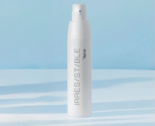 A white, bag-in-bottle airless pump bottle labeled IRRESISTIBLE by Aptar stands upright on a light blue and white surface with a soft blue background.