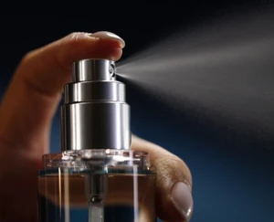 A close-up shows a hand pressing a silver spray pump on a clear bottle as a fine mist disperses horizontally against a dark blue background.