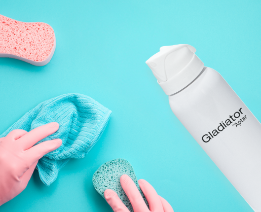 A person wearing pink gloves cleans a blue surface with a blue cloth and green sponge. On the right, a white aerosol can labeled Gladiator, an ergonomically-designed aerosol actuator, next to a pink sponge.