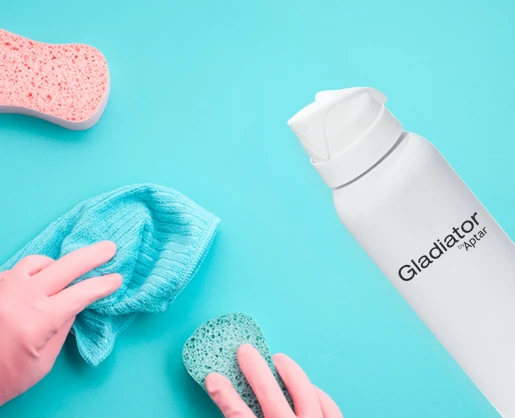 A person wearing pink gloves cleans a blue surface with a blue cloth and green sponge. On the right, a white aerosol can labeled Gladiator, an ergonomically-designed aerosol actuator, next to a pink sponge.