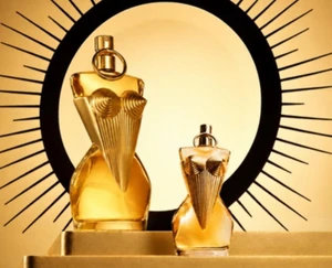 One Jean Paul gaultier Divine Fragrance bottle stands next to a paper fragrance sampling version that is the exact replica of the bottle. They are displayed on a gold pedstal in front of a decorative sunburts backdrop, highlighting how consistent branding is possible for sampling and the full size product.