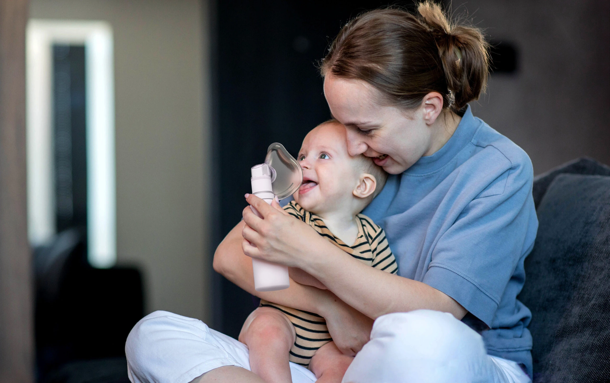 Mother in a blue shirt holds young infant in her arms as she places the mask of a PureHale mist dispenser to their face.