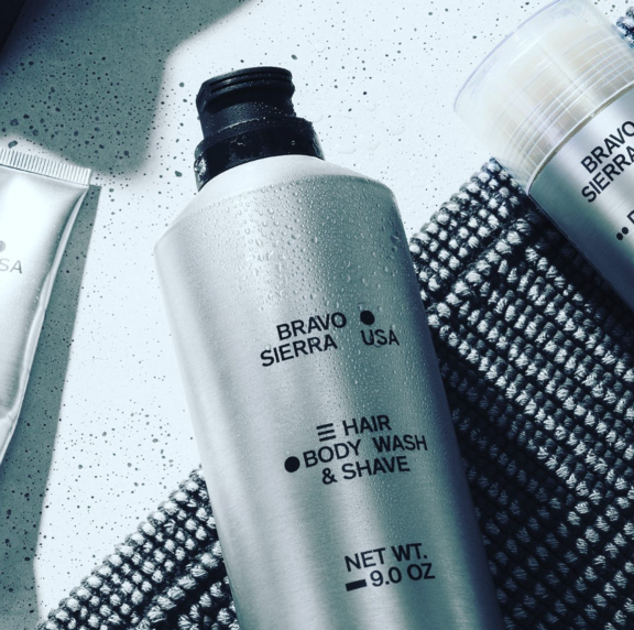A silver bottle with a Contemporary Upright Aerosol Actuator named S20 labeled BRAVO SIERRA USA Hair Body Wash & Shave Net Wt. 9.0 oz sits on a textured gray mat, water droplets on its surface and other grooming products nearby.