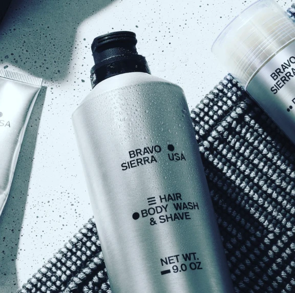 A silver bottle with a Contemporary Upright Aerosol Actuator named S20 labeled BRAVO SIERRA USA Hair Body Wash & Shave Net Wt. 9.0 oz sits on a textured gray mat, water droplets on its surface and other grooming products nearby.