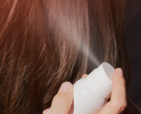 a close up of an aerosol with a white actuator and a white bottle spraying dry shampoo into a woman's hair. The woman's hair is brown.