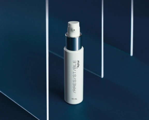 A white airless bottle labeled Irresistible stands upright on a dark blue surface, surrounded by vertical transparent panels. The sleek, modern design features 40 mL airless packaging to preserve the product's freshness.