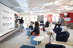 A presenter stands by a large screen displaying the Aptar logo and images, addressing a small group of seated people in a modern, open office space while discussing innovative omnichannel packaging solutions.