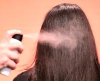 a picture of a hand spraying glitter into long brown hair from behind. The background is a light color pink and the glitter is shimmering in the light..