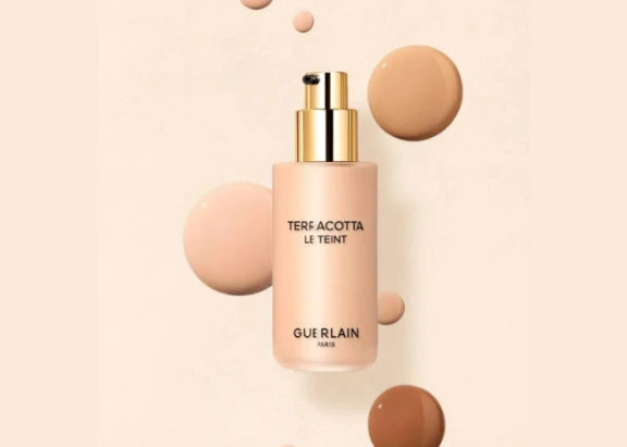 A bottle of Guerlain Terracotta Le Teint foundation featuring Evolux, a chic and premium cosmetic pump, surrounded by foundation swatches of various shades on a light beige background.