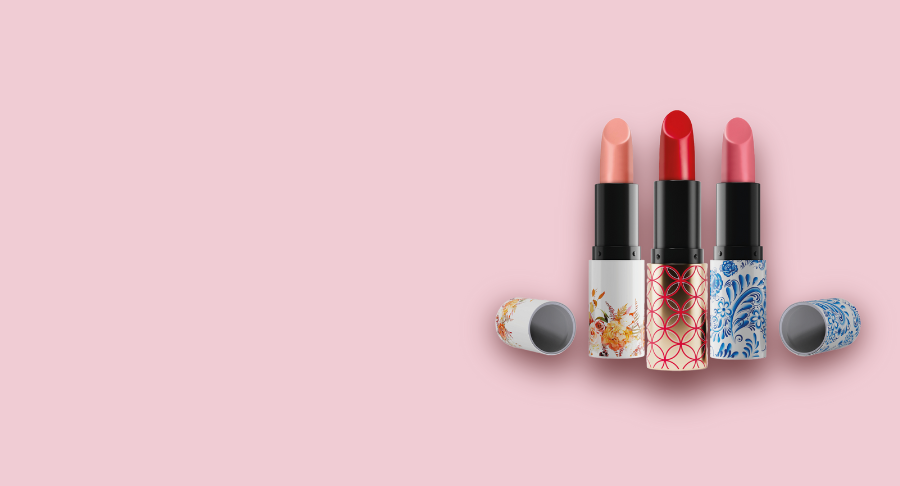 Three lipsticks in refillable lipstick packaging, housed in floral and patterned tubes, stand upright on a pink background. Their caps are placed beside each one. The lipstick shades are nude, red, and pink from left to right.