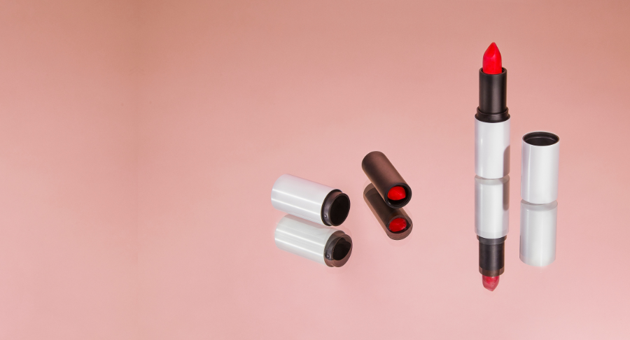 A red lipstick in refillable lipstick packaging stands upright on a reflective pink surface, its cap and reflection beside it. Another closed lipstick tube lies nearby.