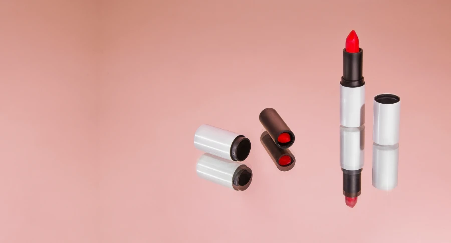 A red lipstick in refillable lipstick packaging stands upright on a reflective pink surface, its cap and reflection beside it. Another closed lipstick tube lies nearby.