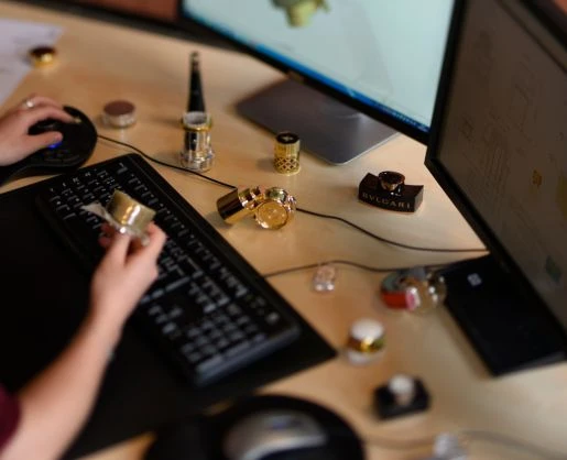 A person works at a computer desk with two monitors, surrounded fragrance caps and other branded components of custom primary beauty packaging—scattered around the keyboard and mouse.