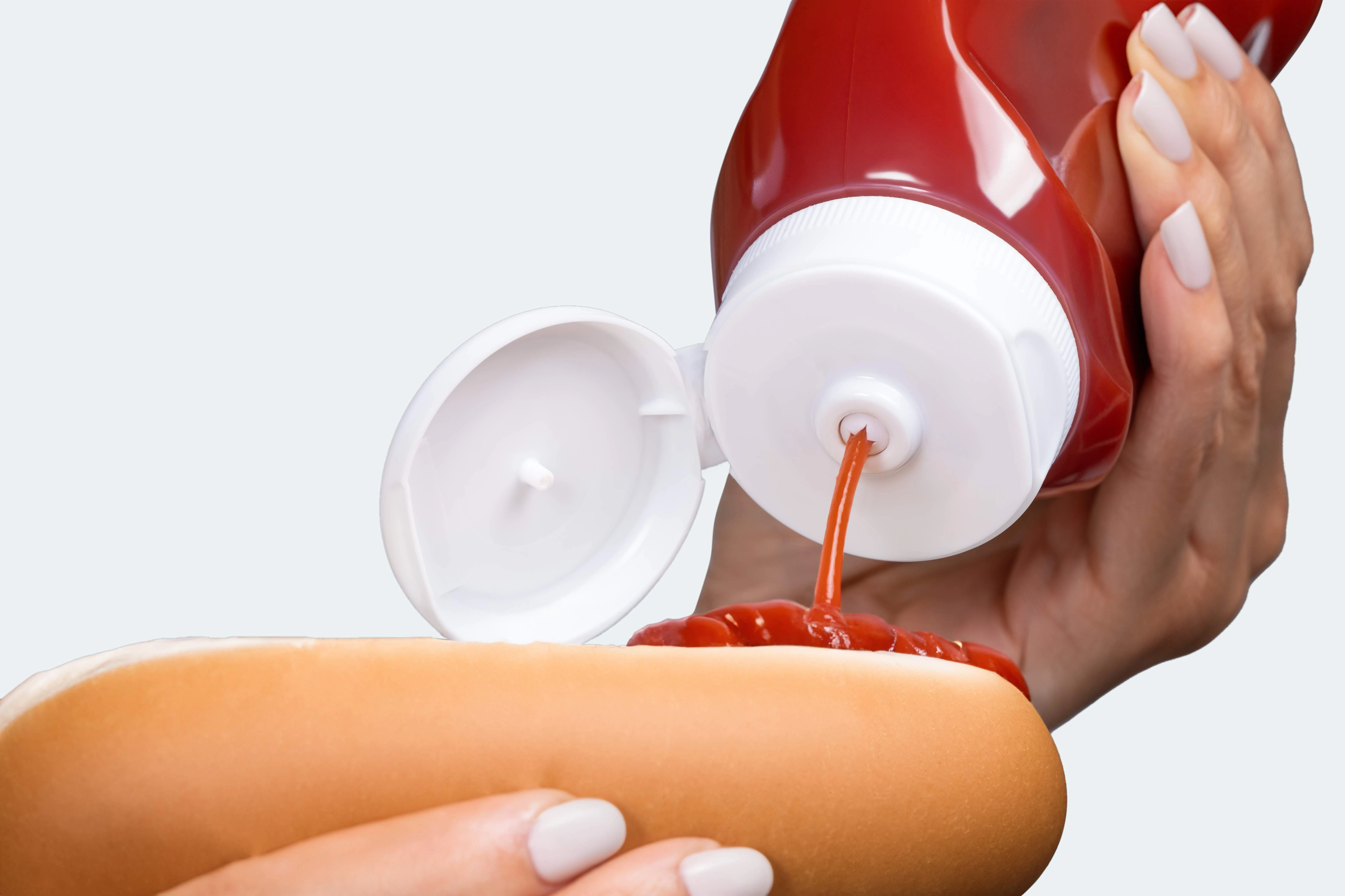 A person squeezing kethcup from an inverted ketchup bottle onto a hot dog