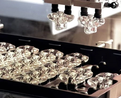 A close-up of a manufacturing machine processing multiple clear glass fragrance caps organized in a tray, with robotic arms above placing or assembling the items—showcasing the precision involved in producing custom primary beauty packaging.