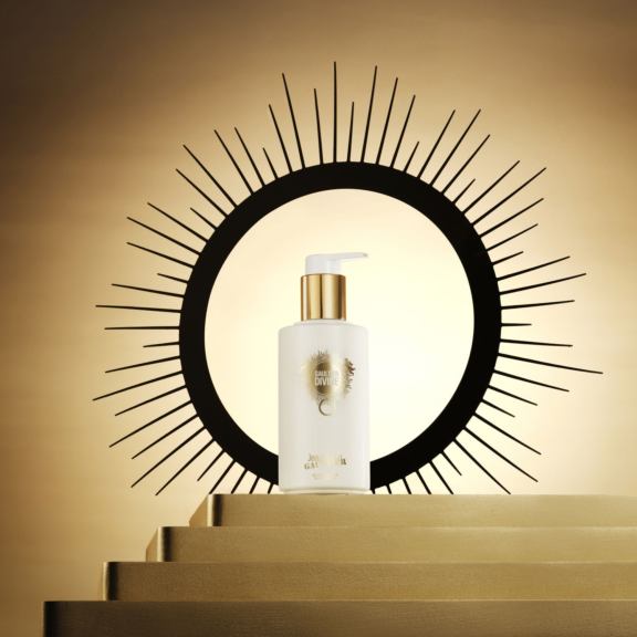 A white and gold Generous Dosage Cosmetic Pump labeled Satine  bottle stands on gold steps, framed by a black circle with radiating lines against a golden background. The arrangement gives the appearance of a glowing sunburst behind the bottle.