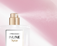 A close-up of a perfume bottle labeled PRECIOUS INUNE by Aptar, featuring a prestige fragrance pump with a shiny gold nozzle, spraying a fine mist against a soft pink background.