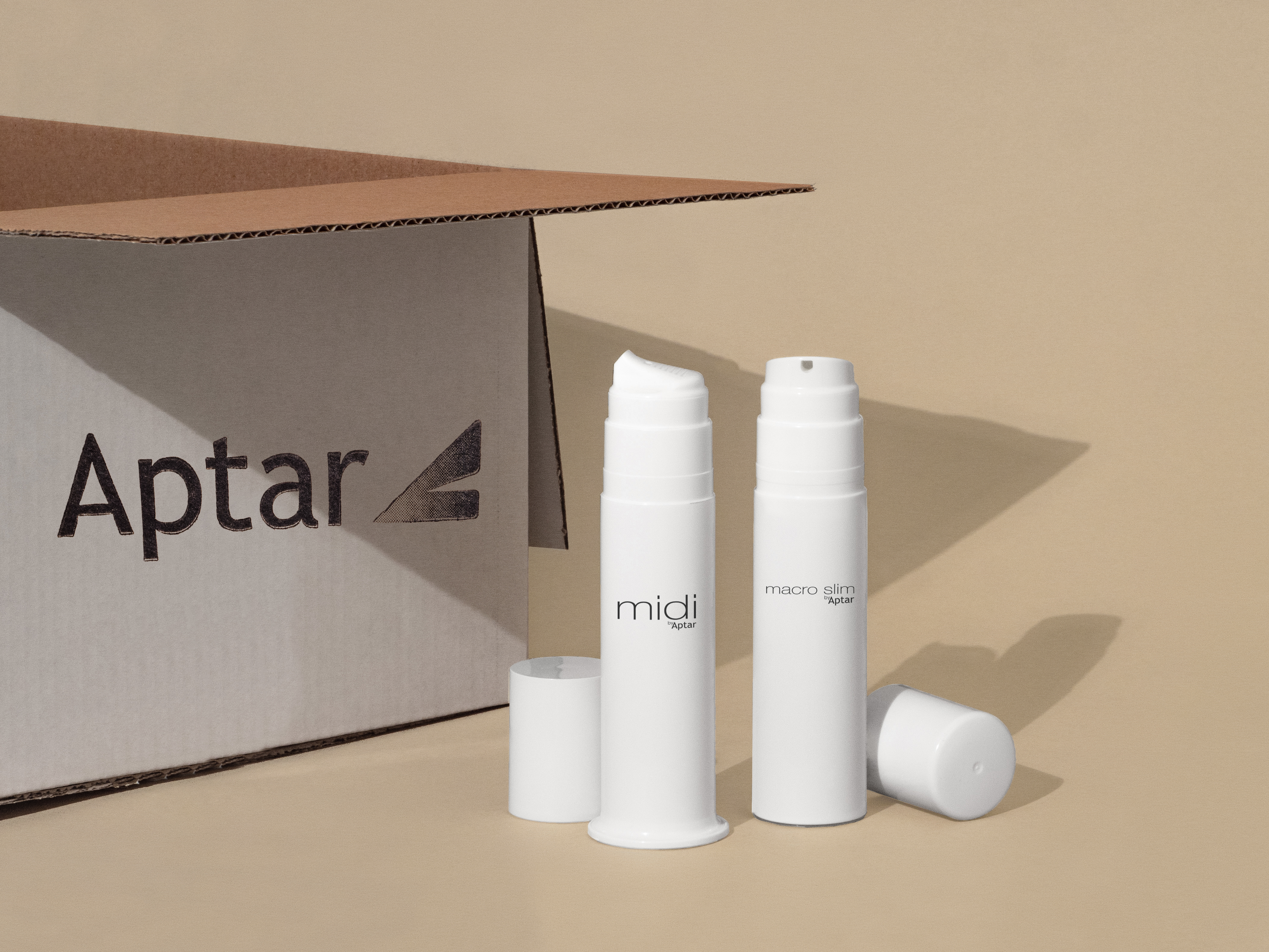 Two white bottom fill airless packaging bottles labeled midi and macro slim stand upright next to an open cardboard box with the word Aptar printed on it, against a beige background.