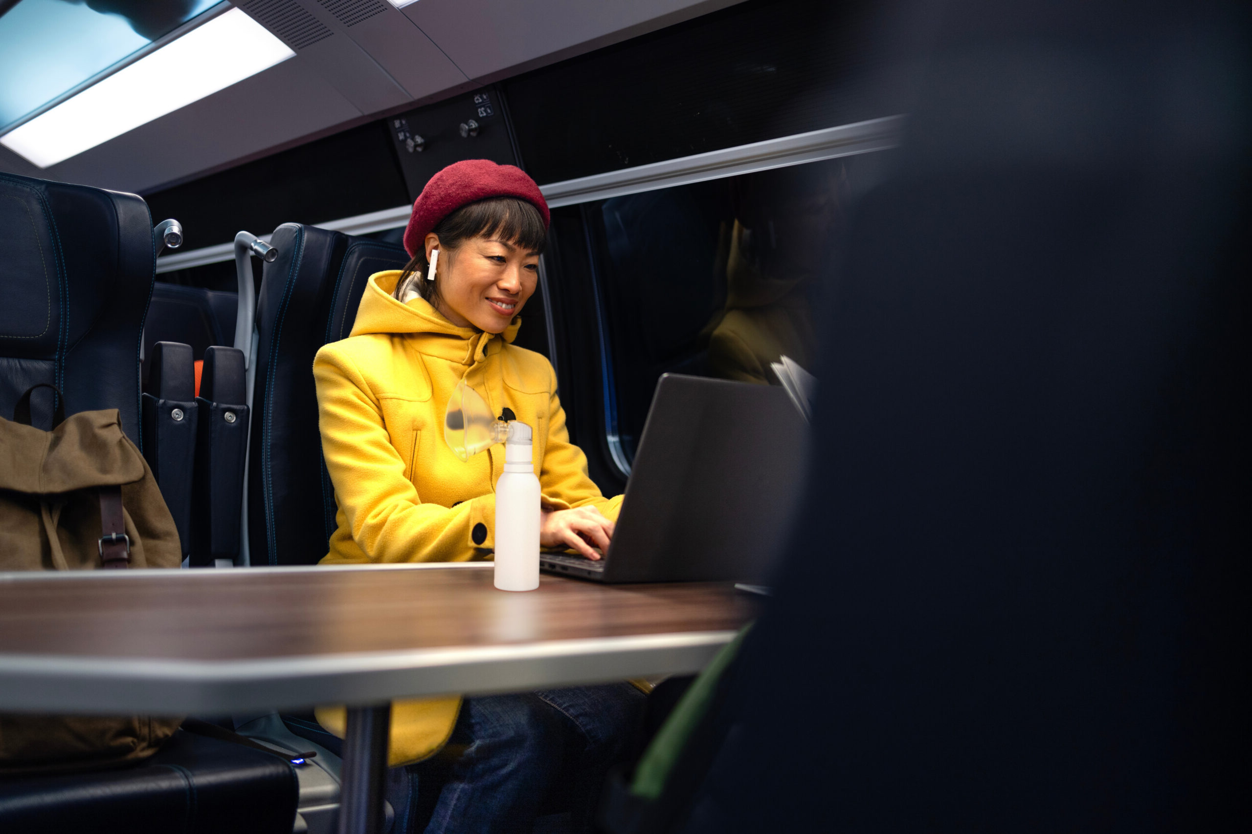 Woman in a yellow coat and red hat types on a computer on a train with a white PureHale fine mist dispenser beside her.