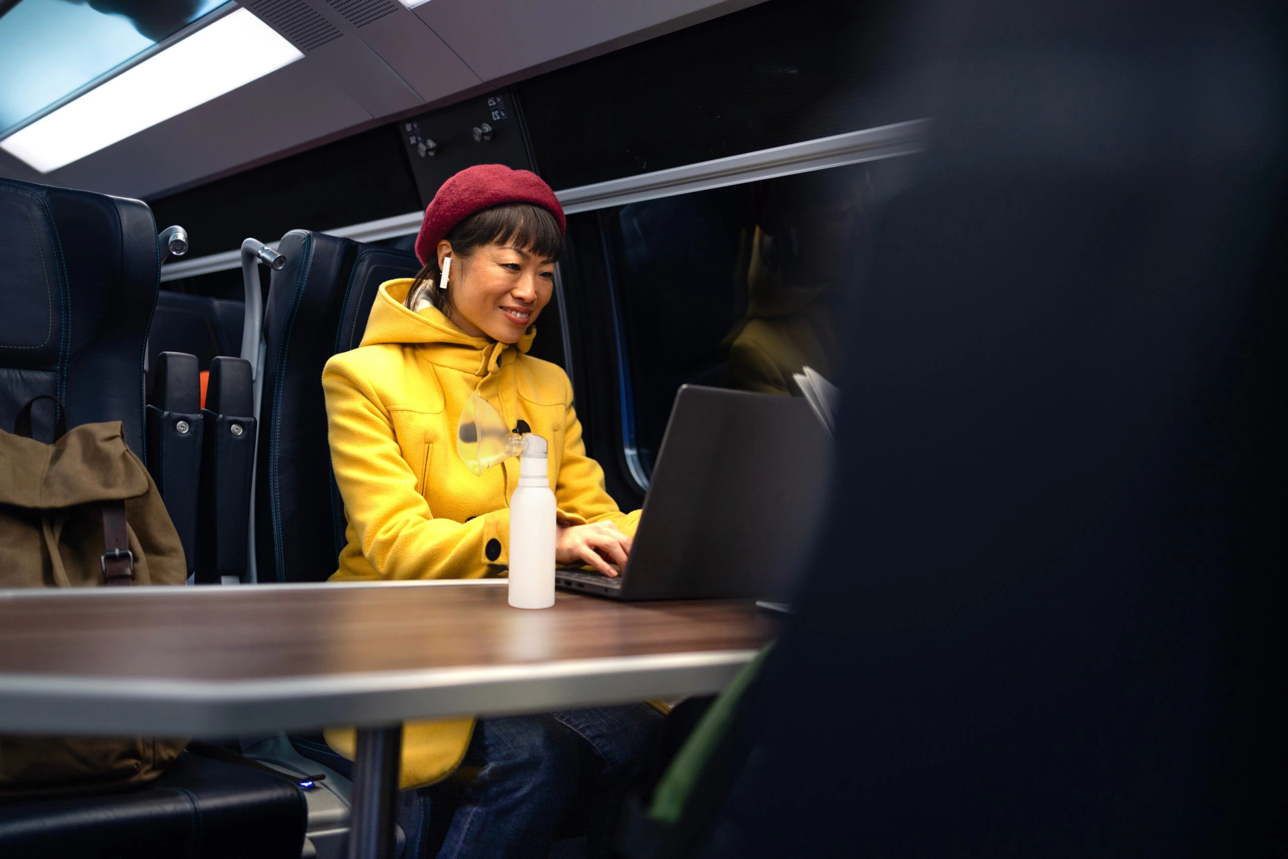 Woman in a yellow coat and red hat types on a computer on a train with a white PureHale fine mist dispenser beside her.
