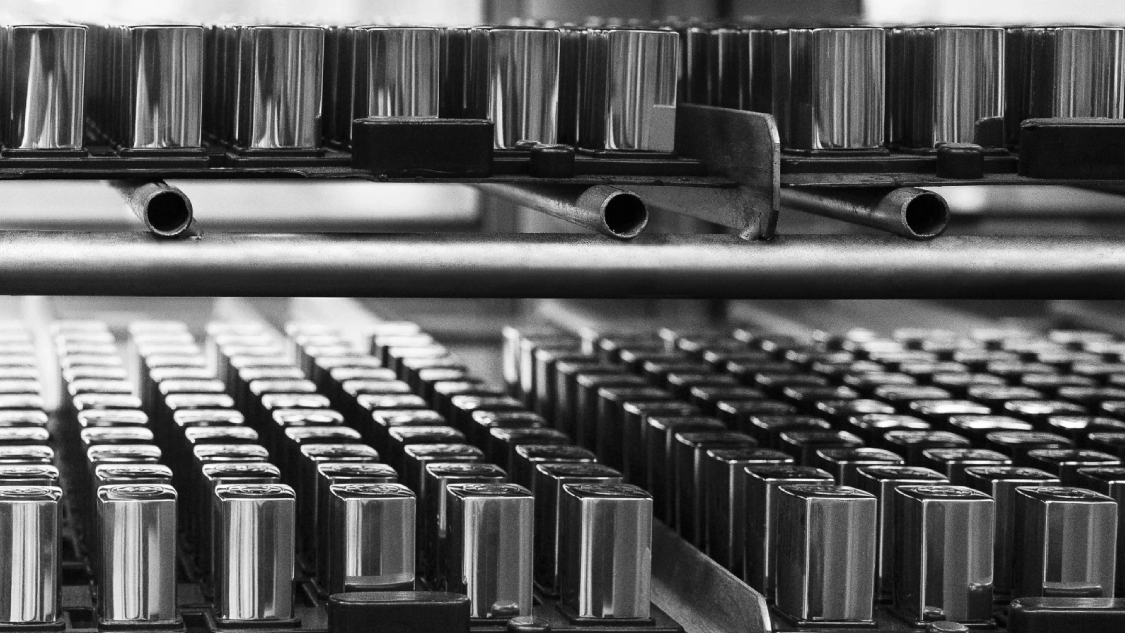 Rows of identical, shiny rectangular metal lipstick cases are stacked on shelves, viewed from a low angle in black and white, evoking the precision and repetition found at a custom cosmetics packaging manufacturer.