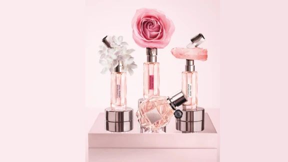 Three elegant perfume bottles with silver caps are displayed on shiny pedestals. Each bottle, featuring a unique fragrance applicator named Note, is topped with white flowers, a pink rose, or a pink crystal. A fourth bottle lies in front. The products are labeled Flowerbomb showing Note on the Victor&Rolf fragrance.