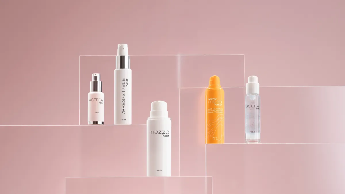 Five airless packaging components - in various shapes and sizes are displayed on clear acrylic stands against a soft pink background. The bottles are mostly white, with one orange and one clear. Each bottle has the product name on it, as a curated selection manufactured by Aptar Beauty.