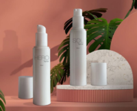 Two white bottles of versatile airless packaging, labeled HERO Aptar and SOL Aptar, stand on a pink surface with a marble slab, green leaves, and a peach-colored background. One bottle cap is off and lying nearby.